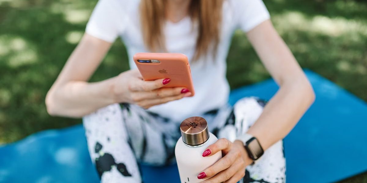 Woman using smartphone while holding bottle on yoga mat outdoors.
