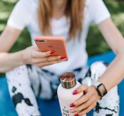 Woman using smartphone while holding bottle on yoga mat outdoors.